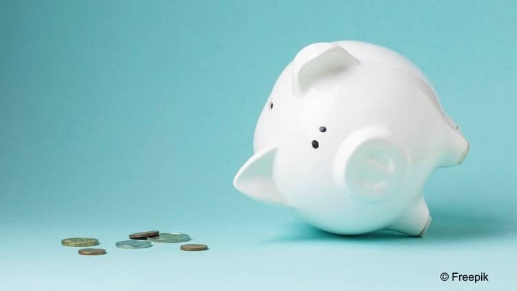 Weißes Sparschwein, welches auf der Seite liegt, als wären die Finanzen in Schieflage geraten. Daneben ein paar Euromünzen vor hellblauem Hintergrund.